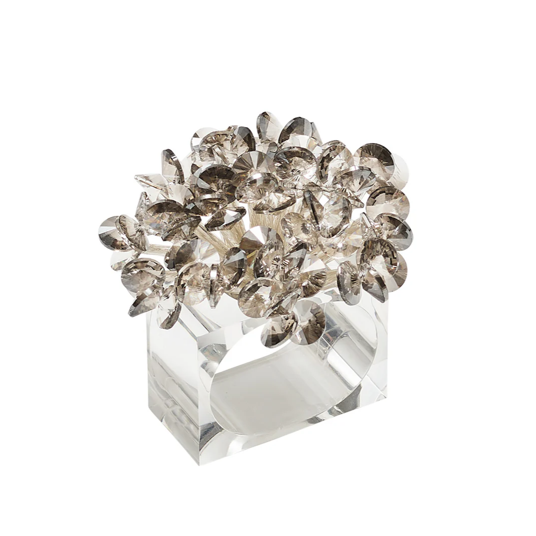 The Aster Napkin Ring Set of 4 by Kim Seybert features clear, rectangular beading with a cluster of faceted, smoky crystal petals on top, bringing an elegant, sparkling floral accent to your table decor.
