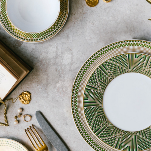 Elegant table setting with Bernardaud Augusta dinnerware—white plates with green and gold geometric patterns—paired with gold cutlery, an open book, jewelry, and gold coins on a light stone surface.