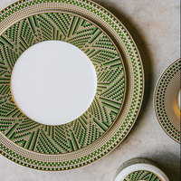 The Augusta Dinnerware by Bernardaud includes three ceramic plates and a bowl adorned with green, gold, and white geometric Art Deco patterns—featuring squares and zigzags—arranged on a textured beige surface.