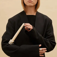A person in a black coat holds an ivory candle in the Babylone Candlestick Holder by Christofle, its silver-plated floral design gleaming. Only the lower face and ring-adorned hands are visible—a striking interior decor moment.