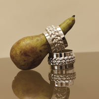 Two silver-plated Babylon Silver Plated Napkin Ring Set 4 rings with a braided design, one encircling a pear and the other resting on a reflective surface.