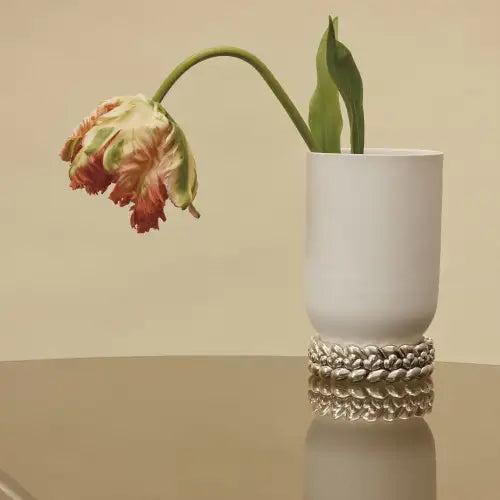 A single red and yellow tulip droops over the edge of a tall Babylone Vase by Christofle, handmade in France. The porcelain vase features a textured band at its base and sits on a reflective surface against a beige background.