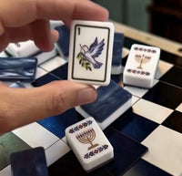 A hand holds a white acrylic tile from the Mahjong Tile Set - Brilliant Nights Hanukkah in Gift Box, featuring a dove with an olive branch, with other tiles depicting menorahs scattered on a checkered board.