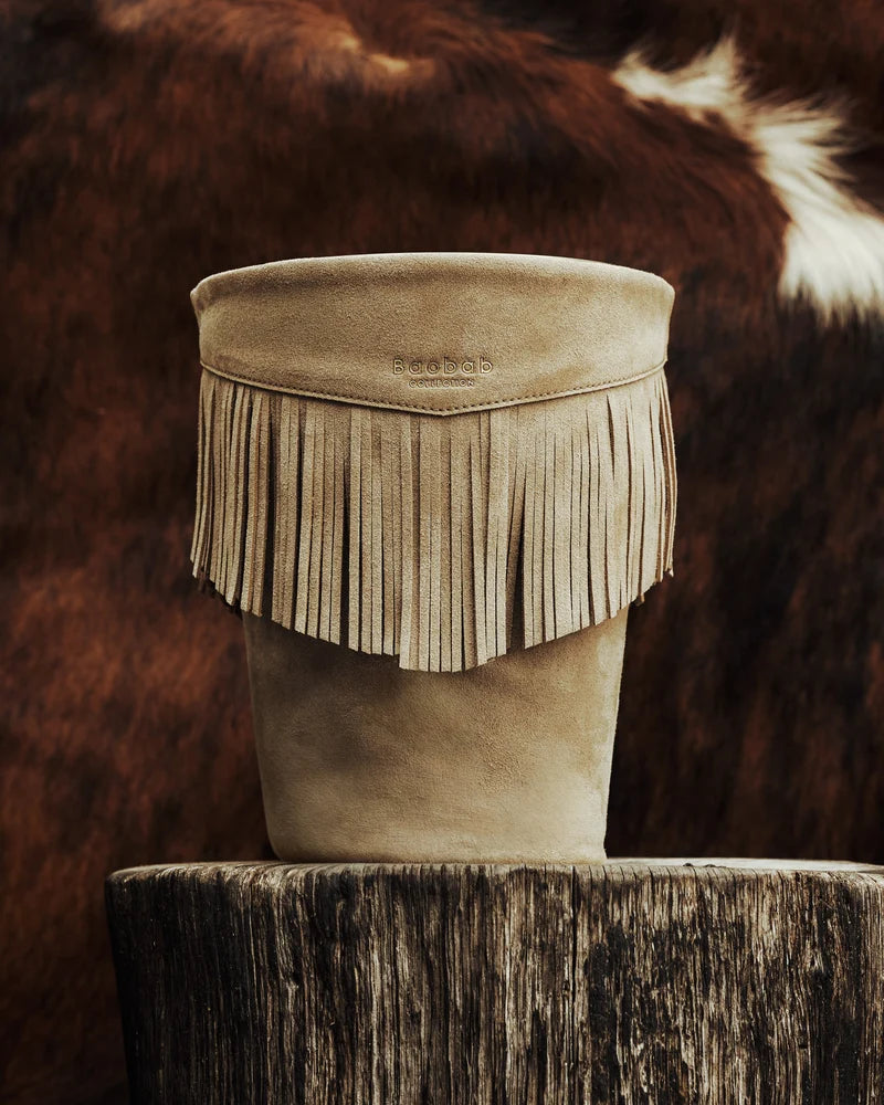 A mineral taupe leather Calamity Jane Candle - The Plains holder with fringe detail, resting on a tree stump.