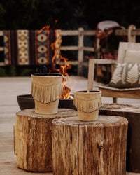 Two fringed mineral taupe Baobab Calamity Jane Candle - The Plains candles sit on tree stumps in front of a fire pit.