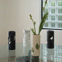 Four Pietra Marble Vases by Greg Natale, in black, white, and beige marble with biomorphic design, are displayed on a glass table. One beige vase holds green stems. A glass block window is visible in the background.