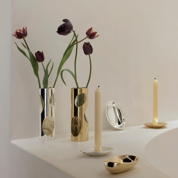 Two Pietra Plated Vases by Greg Natale, featuring cutout circles in a biomorphic design, hold dark tulips. Two tall candles in gold holders and a small gold dish sit nearby on a white surface with a curved edge against an off-white background.