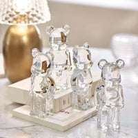 Four Baccarat Bear Brick crystal figurines are showcased on stacked books atop a marble surface, with a gold-accented lamp and decorative objects softly glowing in the background.