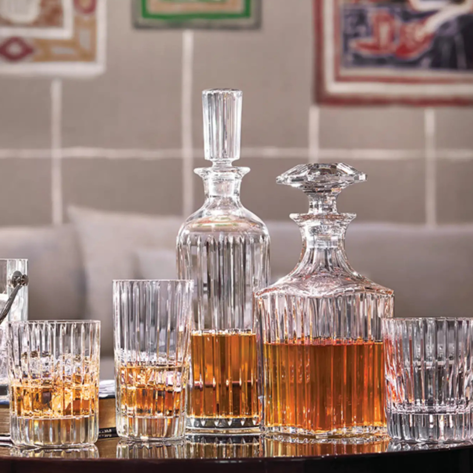 The Baccarat Harmonie Barware Set of 2 by Baccarat features two crystal decanters, surrounded by lead crystal shot glasses and modern highball glass, all arranged on a dark reflective surface with framed art in the background.
