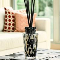 The Baobab Totem Pearls Black Diffuser, featuring a glazed ceramic cap and patterned glass bottle, sits on a dark table. In the background, a white sofa with patterned cushions is softly lit.