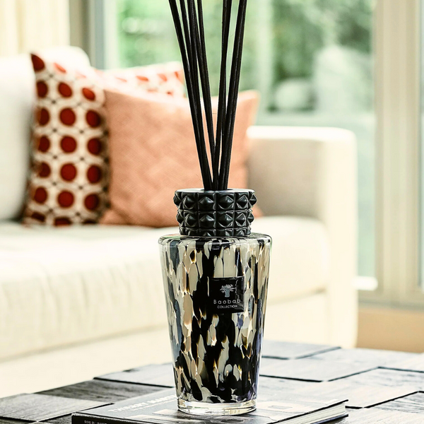 The Baobab Totem Pearls Black Diffuser, featuring a glazed ceramic cap and patterned glass bottle, sits on a dark table. In the background, a white sofa with patterned cushions is softly lit.