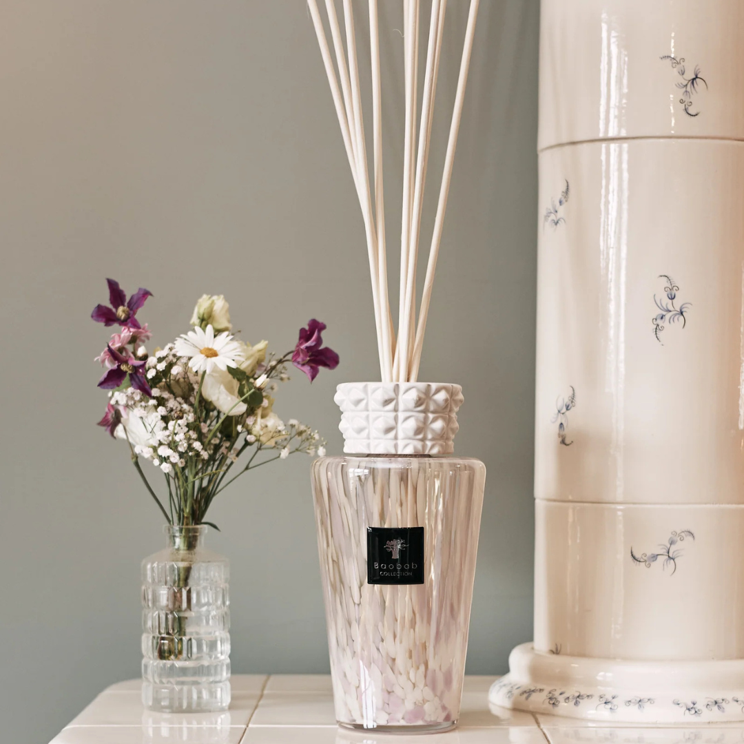 The Baobab Totem Pearls White Diffuser fills the room with a gentle fragrance, its long reeds set beside wildflowers. Behind them is a cream ceramic piece with blue floral patterns.