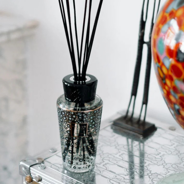 The Baobab Pearls Diffuser, featuring clear liquid and black reeds, is displayed on a glass table next to a colorful patterned vase.