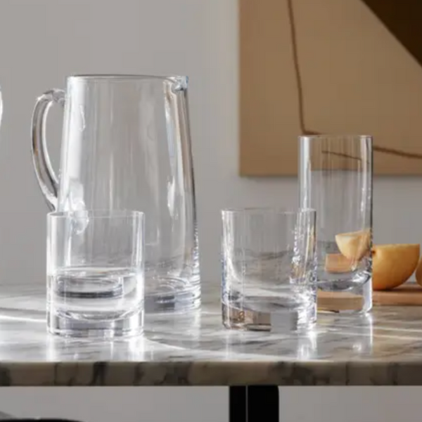 An LSA Bar Drinkware glass pitcher and three classic glasses sit on a marble table; one glass holds two lemon slices, while more lemon pieces rest on a wooden cutting board in the background.