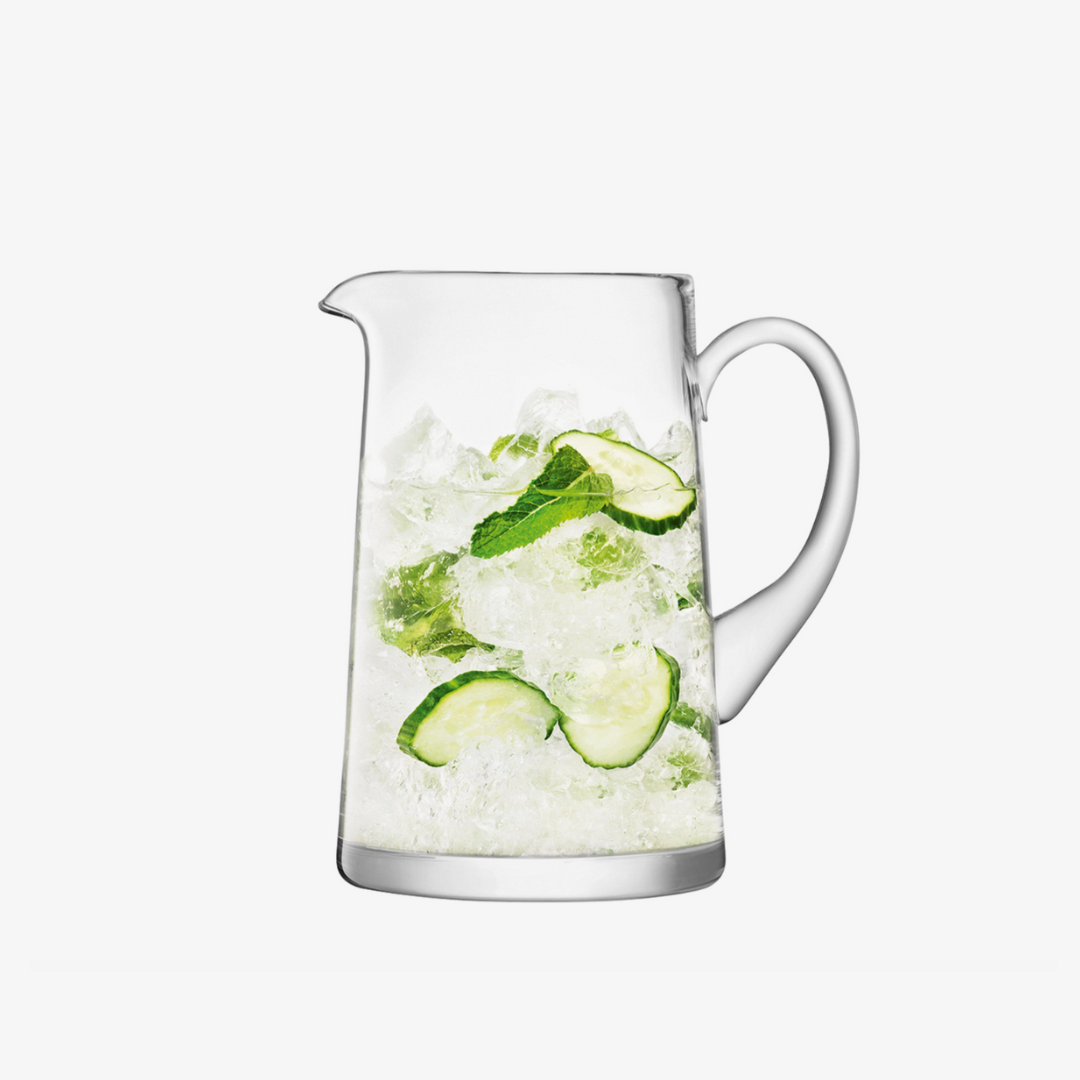 The LSA Bar Tapered Pitcher, a mouth-blown glass jug, is filled with ice, cucumber slices, and mint leaves and set against a white background.