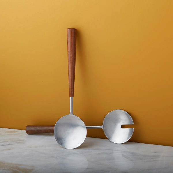 The Bumi Salad Serving Set by Be Home features two modern serving spoons with polished metal scoops and handcrafted rosewood handles, displayed on marble against a golden yellow background.