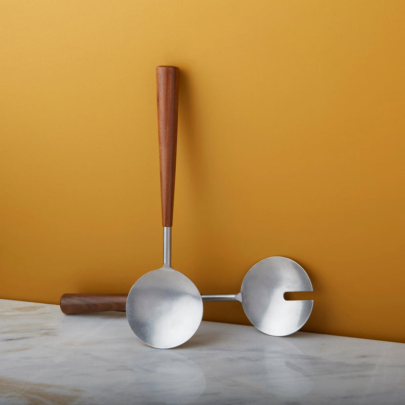 The Bumi Salad Serving Set by Be Home features two modern serving spoons with polished metal scoops and handcrafted rosewood handles, displayed on marble against a golden yellow background.