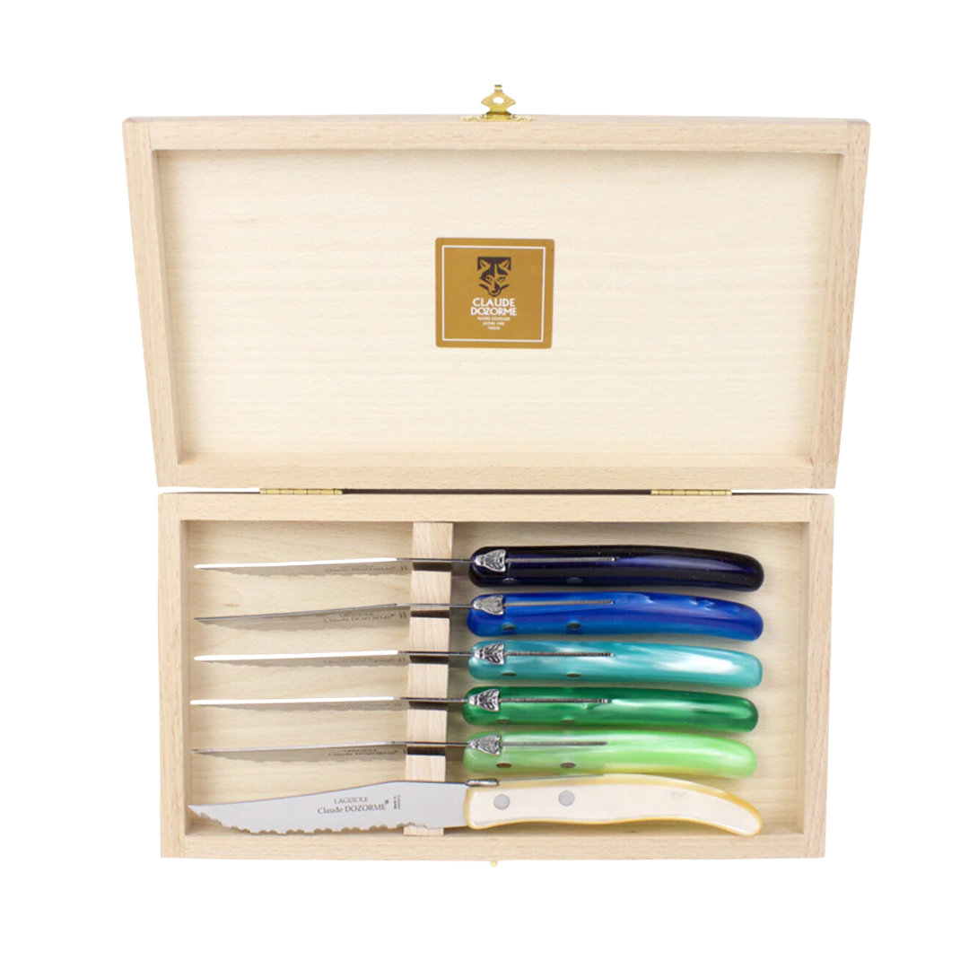 The Claude Dozorme Berlingot Blue & Green Steak Knives Set of 6 comes in a wooden box, each knife featuring stainless steel blades and blue, green, or white handles. The open lid displays the knives and a gold “Made in France” label inside.