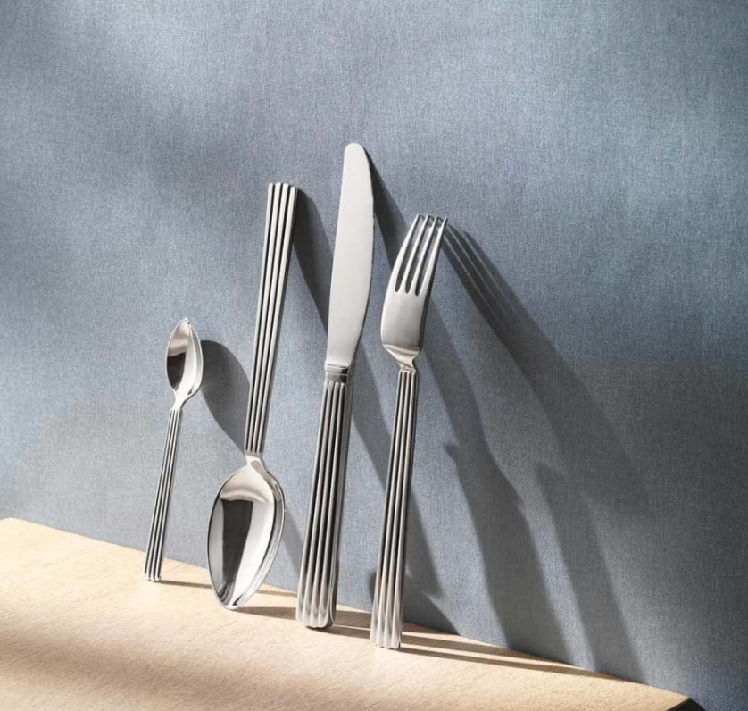 A Georg Jensen Bernadotte Flatware 5 Piece Setting stands upright on a light wooden surface against a textured grey background, each utensil showcasing the signature vertically striped handle design.