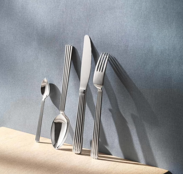 A Georg Jensen Bernadotte Flatware 5 Piece Setting stands upright on a light wooden surface against a textured grey background, each utensil showcasing the signature vertically striped handle design.