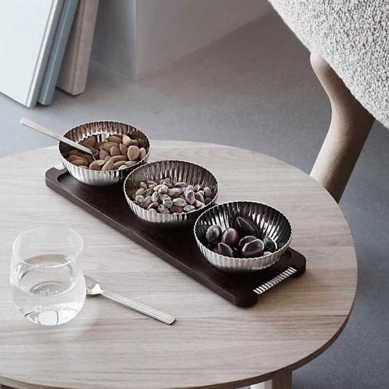 A round light wooden table displays a glass of water with a spoon and the Georg Jensen Bernadotte Triple Bowl Set, filled with almonds, pistachios, and olives. A plush chair and part of the room’s Art Deco floor are visible.