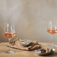 Two glasses of rosé wine on a wooden table with a brown cloth napkin, a smoked oak tray, and the Georg Jensen Bernadotte Triple Bowl Set holding cheese, olives, and pickles against a neutral background.
