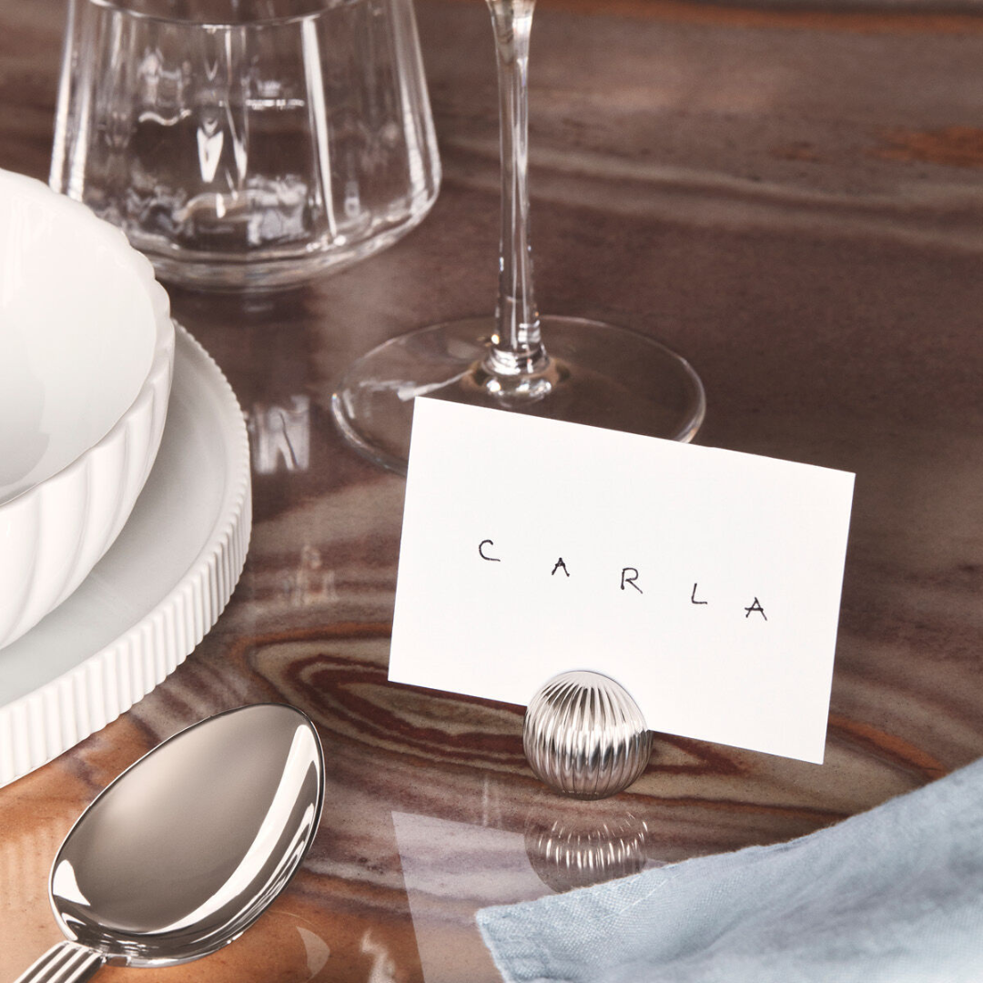 A place card with the name Carla is displayed in a Georg Jensen Bernadotte Table Card Holder on a marbled table next to a spoon, white bowl on a plate, glass, and wine glass.
