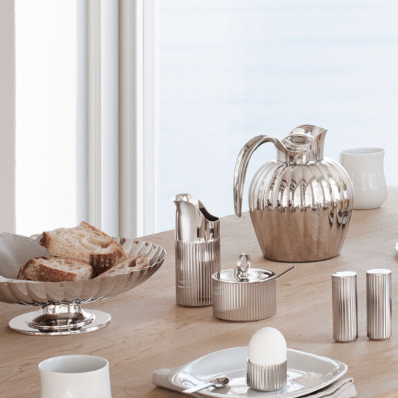 A wooden breakfast table by a window is set with stainless steel silverware, including a Georg Jensen Bernadotte Sugar Bowl, coffee pot, milk jug, salt and pepper shakers, plus sliced bread, a soft-boiled egg, cup, and plate.