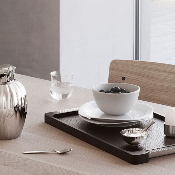 A minimalist breakfast on a light wooden table features the Georg Jensen Bernadotte Wood Tray with a bowl of berries, glass of water, silver pitcher, spoon, small bowl, and soft-boiled egg in an egg cup by the window.