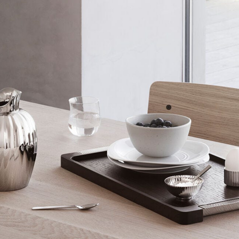 A minimalist breakfast on a light wooden table features the Georg Jensen Bernadotte Wood Tray with a bowl of berries, glass of water, silver pitcher, spoon, small bowl, and soft-boiled egg in an egg cup by the window.