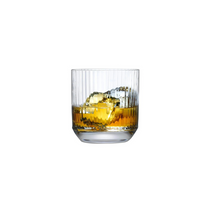 A Big Top Barware Set of 4 crystal glass by Nude Glassware, featuring vertical ridges, is filled with amber liquid and ice cubes against a white background.