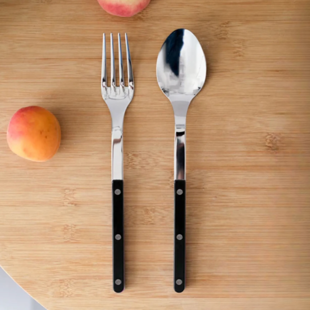 A Sabre Bistrot Shiny Solid Serving Set with black handles rests on a wooden surface beside two peaches, bringing Parisian flair to the scene.