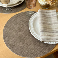 A close-up of a table set with Bodrum Tresse Round Placemat Set of 4, a white scalloped plate, striped linen napkin, ribbed glass, and a vase of cream-colored flowers on a wooden table.