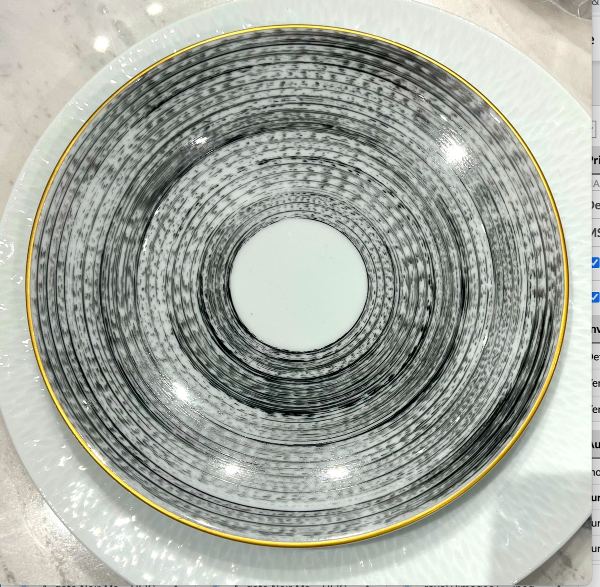 The Marie Daage Agate Dinnerware by Marie Daâge features a round ceramic plate with concentric black and white circles and a gold rim, set atop a larger textured white plate on a light marble surface.