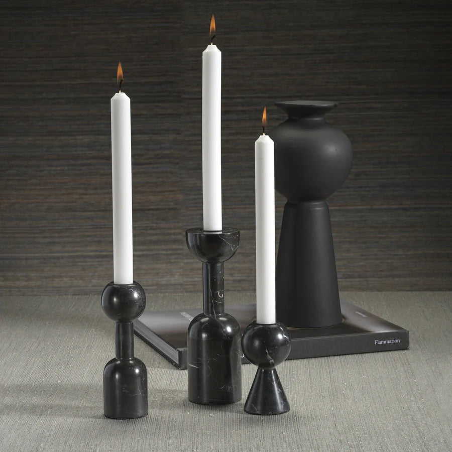 Three Kym Black Candle Holders by Zodax, each holding a white taper candle, are arranged on a light gray surface. A matte black vase on a closed book against a textured gray wall enhances their modern, elegant appeal.