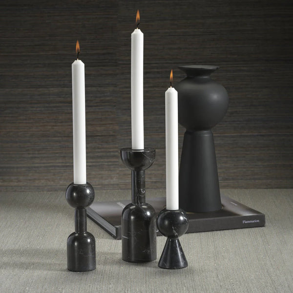 Three Kym Black Candle Holders by Zodax, each holding a white taper candle, are arranged on a light gray surface. A matte black vase on a closed book against a textured gray wall enhances their modern, elegant appeal.