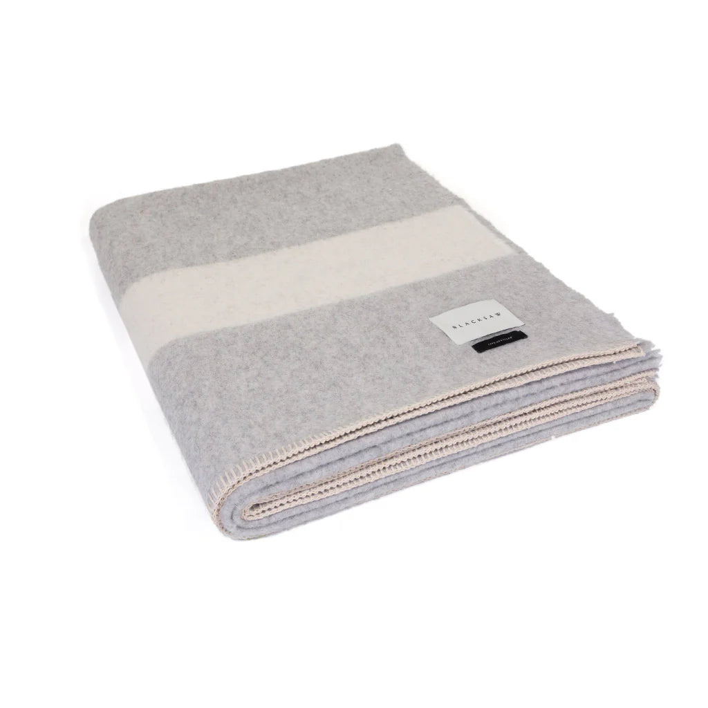 The Siempre Recycled Alpaca Throw by Blacksaw is a heavyweight blanket with wide gray and off-white stripes, beige-stitched edges, and a rectangular corner label. Made from recycled materials, it’s a cozy fireside essential.