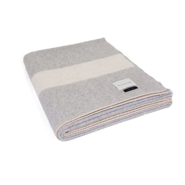 The Siempre Recycled Alpaca Throw by Blacksaw is a heavyweight blanket with wide gray and off-white stripes, beige-stitched edges, and a rectangular corner label. Made from recycled materials, it’s a cozy fireside essential.