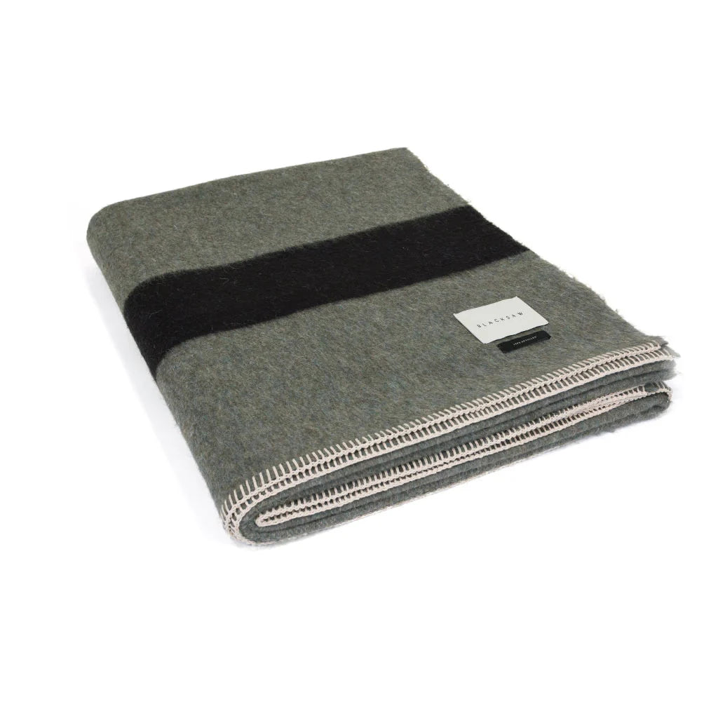 The Siempre Recycled Alpaca Throw by Blacksaw is a heavyweight grey wool blanket with a bold black stripe, white stitched edges, and a rectangular label—a perfect recycled fireside companion.