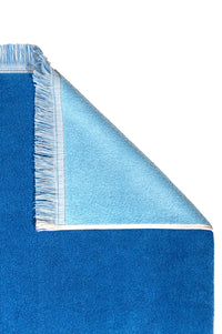The G. McGee Southampton Beach Backgammon is a blue rectangular luxury beach towel with white trim and fringes on one end. A folded top right corner reveals its plush, lighter blue cotton terry underside, set against a white background.