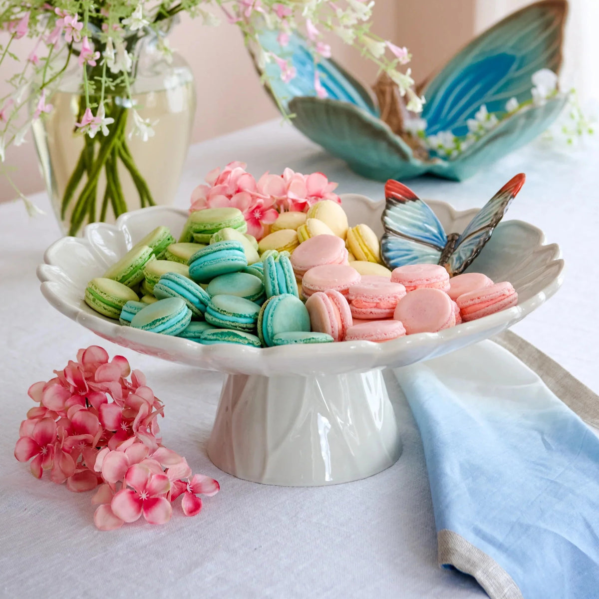 The Vista Alegre Cloudy Butterflies Cake Plate, made of white earthenware and hand painted, beautifully displays pastel macarons in pink, yellow, green, and turquoise with a charming blue butterfly decoration on the side.
