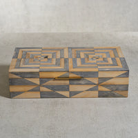 The Zodax Monarch Bamboo Box w/ Brass Trim features a rectangular silhouette adorned with intricate geometric patterns in gray and tan hues, forming concentric diamonds and triangles across the lid and sides, and is elegantly finished with subtle brass trim. Shown here resting on a light textured surface.