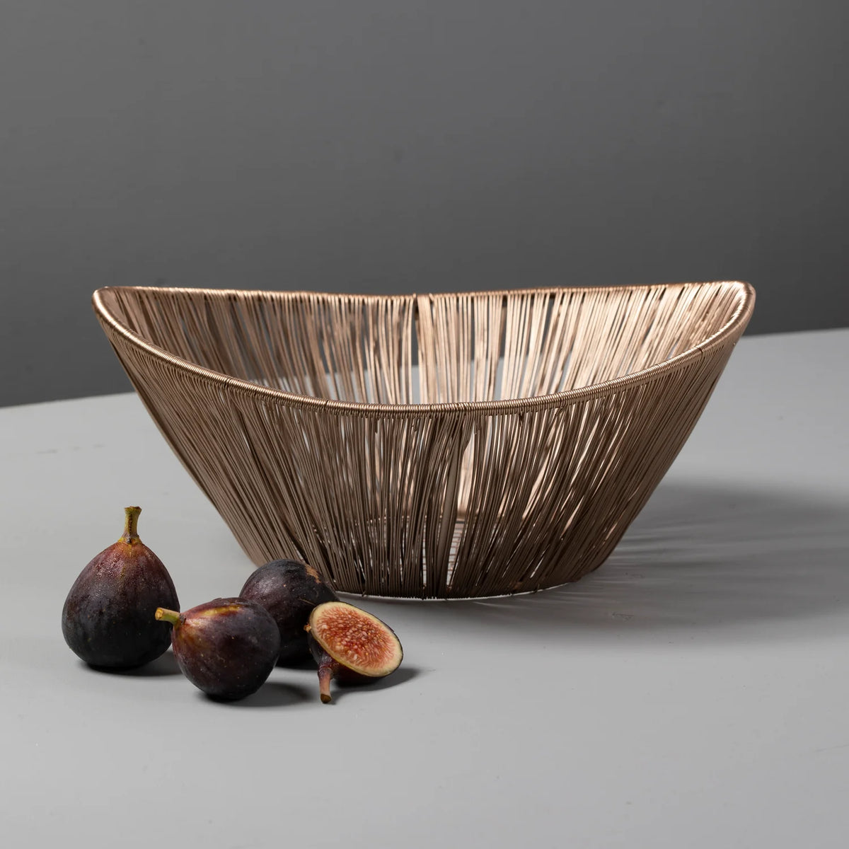 A Be Home Tempo Basket with a metallic finish sits on a gray surface beside four fresh figs, one cut open to reveal its inside. The plain background is also gray.