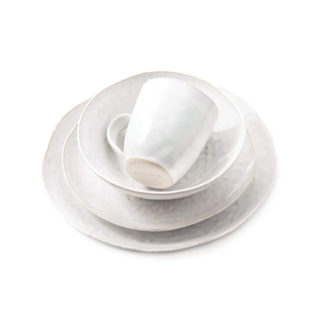 A neatly stacked set of Simon Pearce’s Burlington Dinnerware Cloud is displayed on a white background, including a mug, bowl, salad plate, and an organically edged dinner plate. Each white ceramic piece features tactile uniqueness with subtle freehand pinch-pot design details.