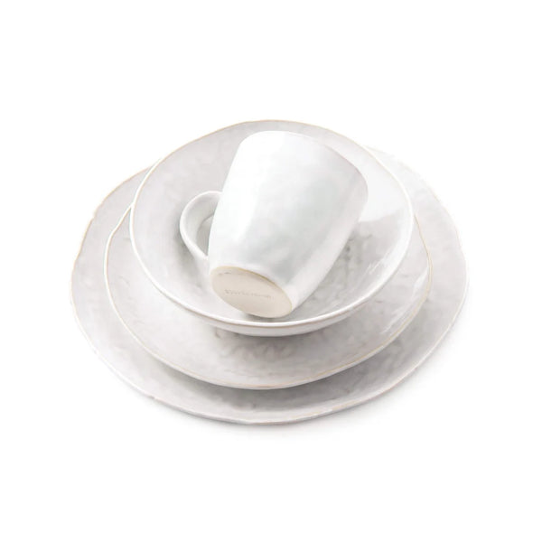 A neatly stacked set of Simon Pearce’s Burlington Dinnerware Cloud is displayed on a white background, including a mug, bowl, salad plate, and an organically edged dinner plate. Each white ceramic piece features tactile uniqueness with subtle freehand pinch-pot design details.