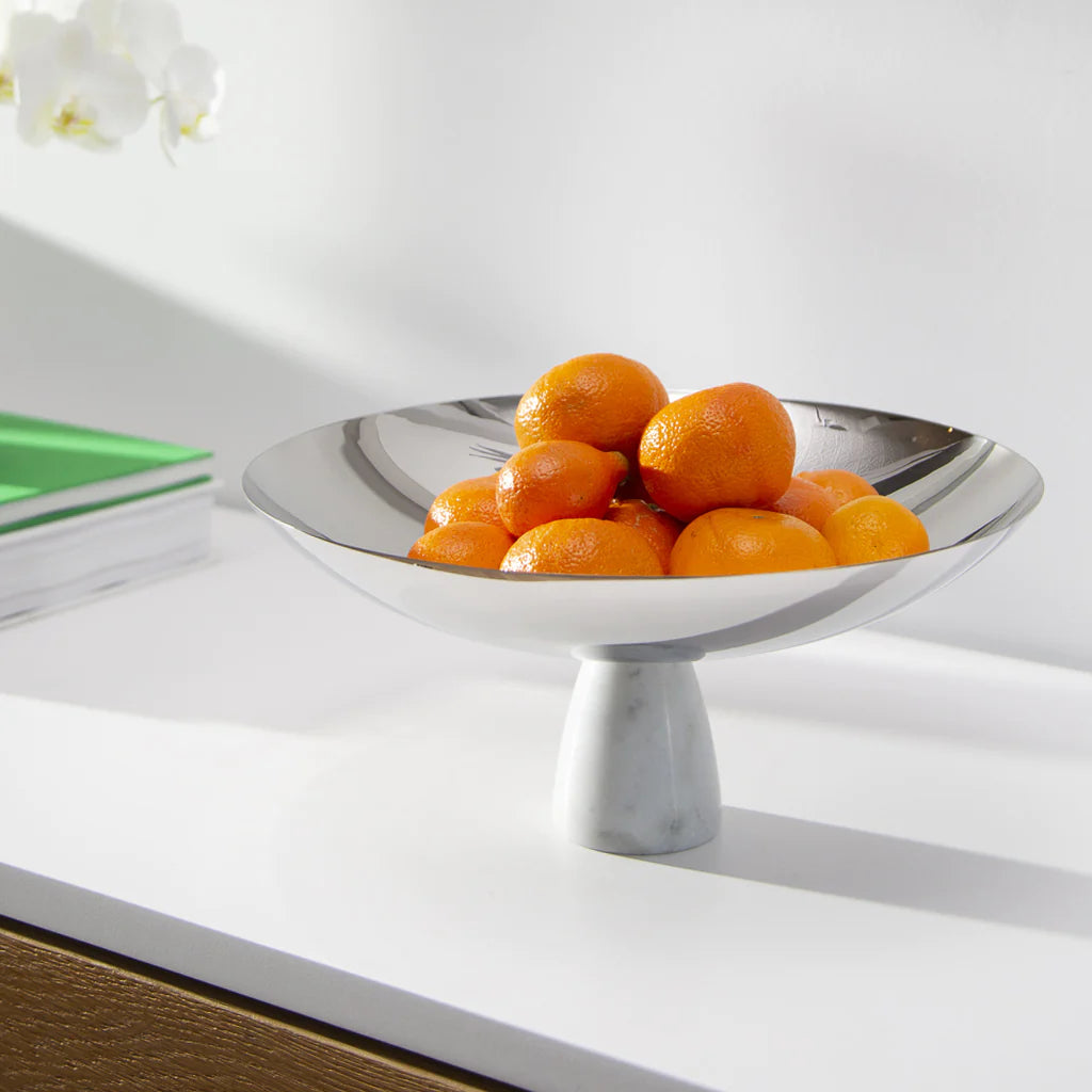 The Coluna Carrara Marble Fruit Bowl by Anna New York displays small oranges on a white countertop, beside green and white books and a partially seen white orchid.