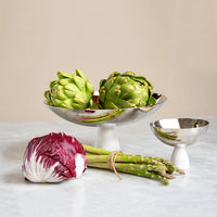 A Coluna Carrara Marble Fruit Bowl by Anna New York—two silver pedestal bowls with green artichokes—sits on a marble table next to twine-tied asparagus and a head of radicchio, all set against a neutral beige background.