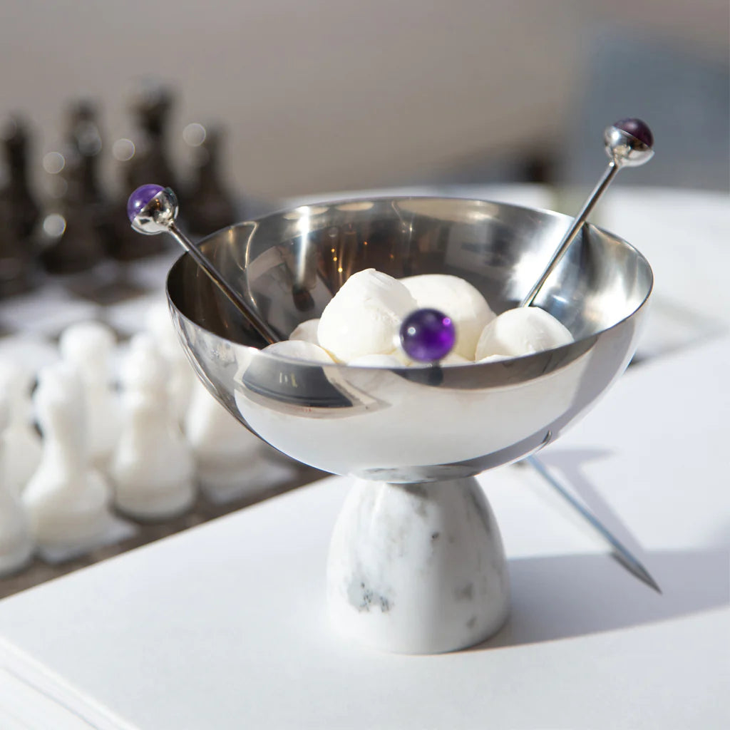 The Anna New York Coluna Carrara Marble Nut Bowl holds white marshmallows, accompanied by two metal picks with purple bead tops. A blurred chessboard with pieces appears in the background.