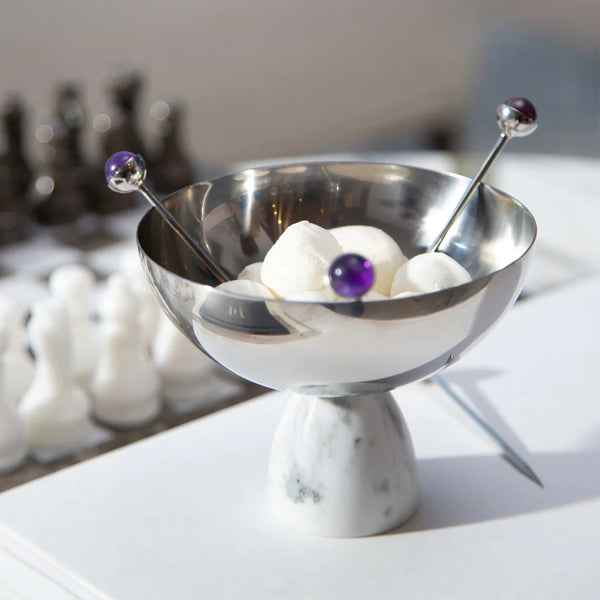 The Anna New York Coluna Carrara Marble Nut Bowl holds white marshmallows, accompanied by two metal picks with purple bead tops. A blurred chessboard with pieces appears in the background.
