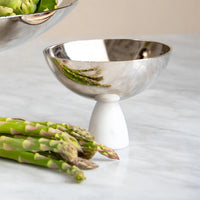 The Anna New York Coluna Carrara Marble Nut Bowl with a white marble base sits on a marble surface beside fresh asparagus, reflecting the green spears in its shiny silver finish.